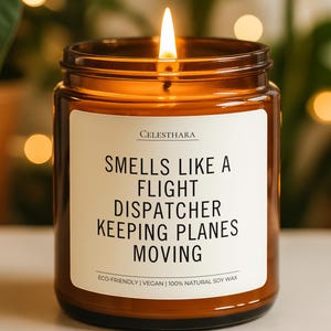 May include: A lit soy wax candle in a brown glass jar. The candle has a white label with the text "SMELLS LIKE A FLIGHT DISPATCHER KEEPING PLANES MOVING". The candle is eco-friendly and vegan.