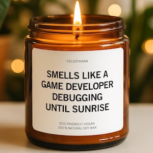 May include: A lit soy wax candle in a brown glass jar. The white label reads "Smells Like a Game Developer Debugging Until Sunrise." The candle is eco-friendly and vegan.