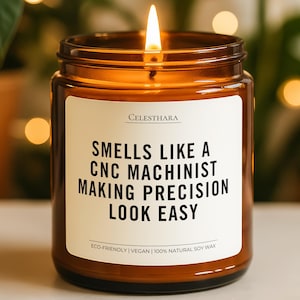 May include: A lit soy wax candle in a brown glass jar. The label reads "SMELLS LIKE A CNC MACHINIST MAKING PRECISION LOOK EASY." The candle is eco-friendly and vegan. The background is blurred with green plants and soft lights.