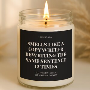 May include: A lit soy wax candle in a clear glass jar. The black label reads "Smells Like a Copywriter Rewriting the Same Sentence 12 Times." The candle is eco-friendly and vegan.