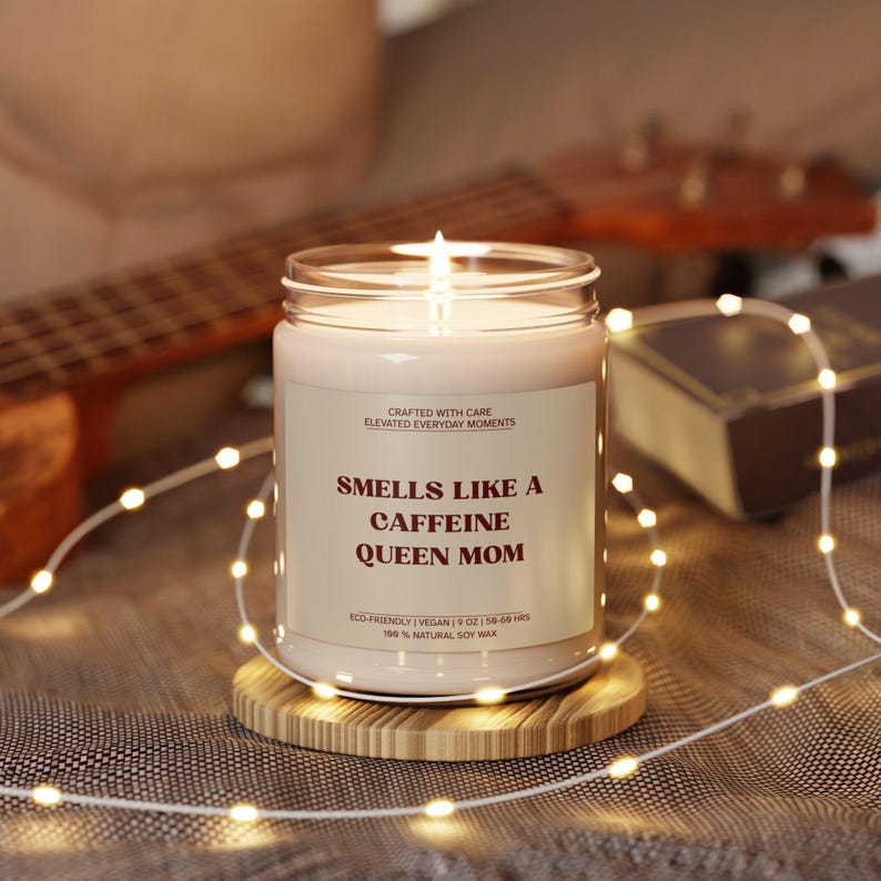 May include: A lit candle in a glass jar with a label that reads "Smells Like a Caffeine Queen Mom." The candle sits on a wooden coaster. The text "Crafted with Care, Elevated Everyday Moments" is also on the label.