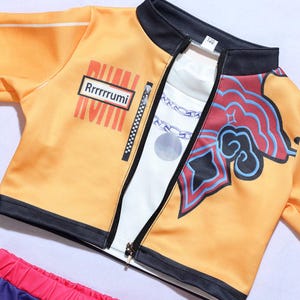 May include: An orange zip-up jacket with black trim, featuring a white inner layer with a chain print. The jacket displays the text Rrrrrrumi on a rectangular patch, a vertical zipper detail, and a red and blue graphic design on the right side.