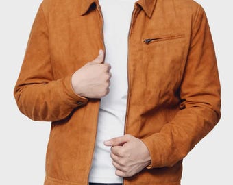 May include: A tan suede jacket with a collared neckline and a front zipper closure. The jacket features a small zippered chest pocket and buttoned cuffs. The model is wearing a white t-shirt underneath. The jacket is a classic style.
