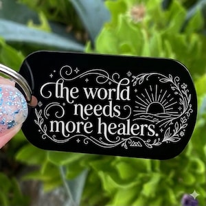 May include: A black rectangular keychain with rounded corners featuring white engraved text that reads, "the world needs more healers." The design includes decorative flourishes, a sun rising over a horizon line, and small star accents.