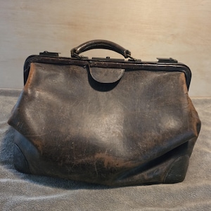 May include: A dark brown leather doctor's bag featuring a structured frame, a single top handle, and a metal clasp closure. The exterior shows visible signs of wear, including surface scratches, scuffs, and faded areas on the leather material.