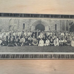 May include: A black and white panoramic photograph of a large group of people posed in front of a stone building with arched windows and a central doorway. The photograph is enclosed in a rectangular frame with a dark, textured, wood-grain pattern.
