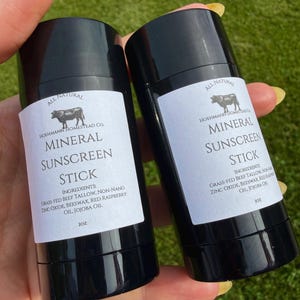 Pode incluir: Dois bastões de protetor solar mineral preto com rótulos brancos. Os rótulos dizem "All Natural", "Mineral Sunscreen Stick" e listam ingredientes como óxido de zinco e óleo de jojoba. Cada bastão tem 28g.