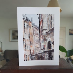 May include: Watercolour painting of a European street scene. The artwork depicts tall buildings with warm, earthy tones, and a narrow street. The painting includes architectural details, street lamps, and a sign that reads "Bistro Ruby".