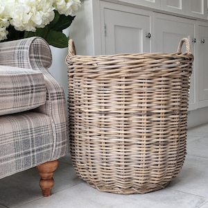 May include: A large, round wicker basket with two handles. The basket is light brown and has a textured surface. The basket is next to a plaid armchair and white cabinets.