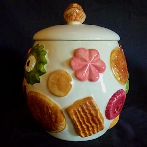 May include: A white ceramic cookie jar with a lid featuring a sculpted handle. The exterior is decorated with various raised, three-dimensional cookie designs, including a pink flower-shaped cookie, a rectangular biscuit, and round brown cookies.