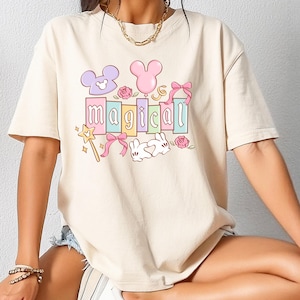 May include: Cream-colored t-shirt with a colorful graphic that says "magical." The design includes Mickey Mouse ears, bows, a wand, and a bunny. The shirt is a relaxed fit.