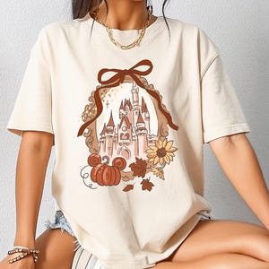 May include: Cream-colored t-shirt featuring a brown castle illustration with a pumpkin, sunflower, and fall leaves. The design is framed by a brown ribbon and a decorative border. The shirt has short sleeves.