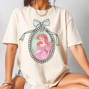 Può includere: T-shirt color crema con un'illustrazione a cartoni animati di una principessa in un abito rosa all'interno di una cornice ovale. La cornice è ornata da un bordo perlato e da un fiocco verde chiaro. La principessa ha i capelli rossi e si trova su uno sfondo verde chiaro.