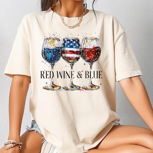 May include: Cream-colored t-shirt featuring a graphic of three wine glasses filled with red, white, and blue liquids, with the text "RED WINE & BLUE" below. The design is reminiscent of the American flag.