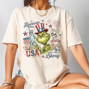 May include: Cream-colored t-shirt featuring a graphic of the Grinch in a patriotic hat, the Statue of Liberty, and a Route 66 hot dog stand. The design includes the text "American Made USA" and "Sweet Land of Liberty."