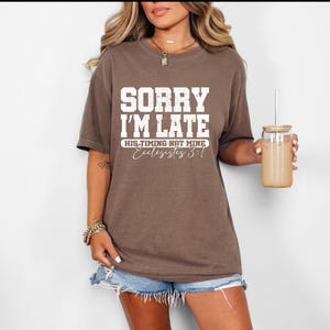 May include: A brown t-shirt with the text "SORRY I'M LATE" in white block letters. Below the text is "HIS TIMING NAT MINE" and "Ecclesiastes 3:1" in a script font. The person is wearing denim shorts and holding a glass with a straw.
