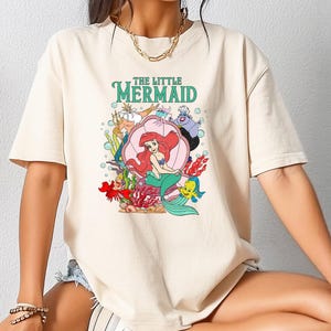 Può includere: T-shirt color crema con grafica colorata de La Sirenetta di Disney. Il design include Ariel, Flounder, Sebastian e Ursula, con il titolo in verde. La maglietta ha una vestibilità casual e oversize.