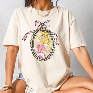 Può includere: T-shirt color crema con un'illustrazione incorniciata di una principessa con capelli biondi e un vestito rosa. La cornice è adornata da un fiocco e un nastro rosa. La maglietta ha maniche corte e una vestibilità ampia.