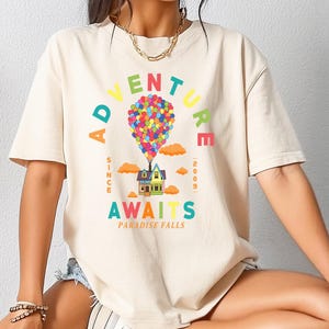 May include: Cream-colored t-shirt featuring a colorful graphic of a house lifted by balloons. The text reads "ADVENTURE AWAITS" with "SINCE 2009" and "PARADISE FALLS" below. The design is in bright, playful colors.