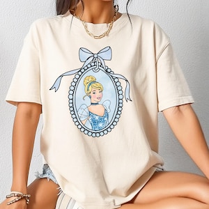 Può includere: T-shirt color crema con un'illustrazione incorniciata di Cenerentola con un vestito blu e un fiocco azzurro. La cornice ovale è delineata da piccole perline bianche. La principessa ha i capelli biondi e indossa un choker.