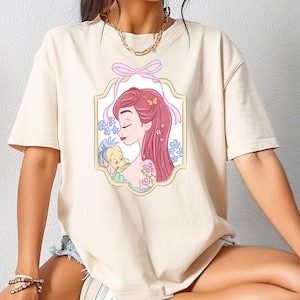 Può includere: T-shirt color crema con un'illustrazione incorniciata di una donna dai capelli rossi e un piccolo pesce giallo. L'illustrazione è circondata da fiori e un fiocco rosa. La maglietta ha maniche corte e una vestibilità ampia.