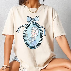 Può includere: T-shirt color crema con un'immagine a cartone animato di una principessa in una cornice ovale blu con un fiocco blu. La principessa ha i capelli bianchi e indossa un vestito blu. La maglietta ha maniche corte.