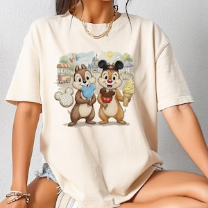 Puede incluir: Camiseta color crema con un gráfico colorido de Chip y Chop sosteniendo golosinas frente a un fondo de parque temático. Chip sostiene un helado azul, mientras que Chop tiene un cono de helado amarillo. También se ve una galleta de Mickey Mouse.