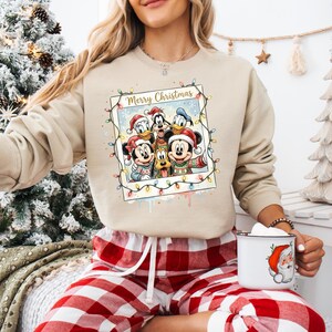 May include: A beige crewneck sweatshirt featuring a graphic print of Mickey Mouse, Minnie Mouse, Donald Duck, Daisy Duck, and Goofy wearing Santa hats. The design is framed like a Polaroid photo with the text Merry Christmas and string lights.
