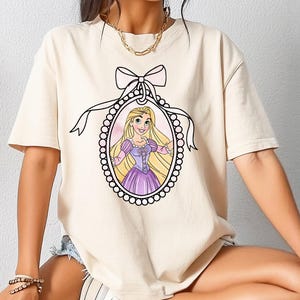 Può includere: T-shirt color crema con un'illustrazione a cartoni animati di una principessa in una cornice decorativa. La cornice è adornata da un fiocco e perle. La principessa ha lunghi capelli biondi e indossa un vestito viola.