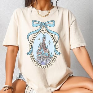 Può includere: T-shirt color crema con un disegno di castello dei cartoni animati all'interno di una cornice ovale, ornata da un fiocco azzurro e abbellimenti a forma di perla. Il castello è raffigurato in rosa, blu e viola, con uno sfondo di cielo azzurro.