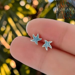 Op de afbeelding: Een paar kleine, lichtblauwe zeester-oorbellen. De oorbellen hebben een zilverkleurige omtrek en worden tussen vingers gehouden tegen een vage groene achtergrond. Een accessoire met nautisch thema.