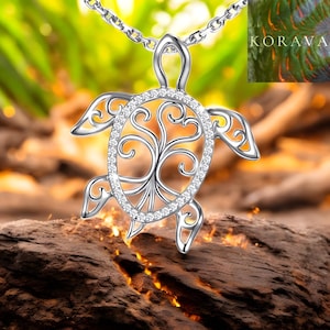 Puede incluir: Un collar con colgante de tortuga plateada. El colgante presenta un diseño de árbol de la vida dentro de un marco con forma de tortuga, adornado con pequeñas gemas transparentes. La cadena del collar también es plateada. La palabra "KORAVA" es visible.
