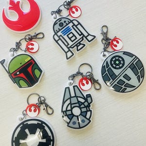May include: A collection of Star Wars-themed keychains. The keychains feature iconic characters and symbols, including R2-D2, Boba Fett, the Millennium Falcon, and the Death Star. Each keychain has a silver clasp and a red and white emblem.
