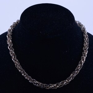 May include: A silver chain necklace with a woven design. The necklace is displayed on a black mannequin.