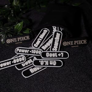 Può includere: Una collezione di segnapunti rettangolari bianchi e neri con scritte come One Piece, Can't Block, Negate, Searcher, Power -1000, Power -5000, Cost +7 e On K.O. Questi piccoli gettoni piatti sono disposti in un mucchio vicino a un sacchetto nero.