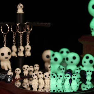 Könnte beinhalten: Eine Sammlung weißer Figuren mit schwarzen Augen und rundem Kopf, einige an Schlüsselanhängern. Die Figuren sind auf einer reflektierenden Oberfläche angeordnet und leuchten teilweise grün. Ein schwarzer Ständer hält Schlüsselanhänger mit denselben Figuren.