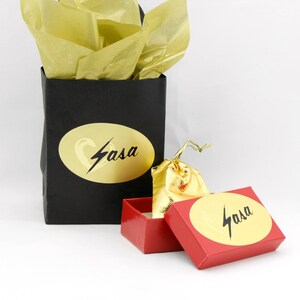 May include: A black gift bag with gold tissue paper, a red box, and a gold drawstring bag. The gift bag and box have a gold oval sticker with the word "Sasa" in black script. The drawstring bag is gold.