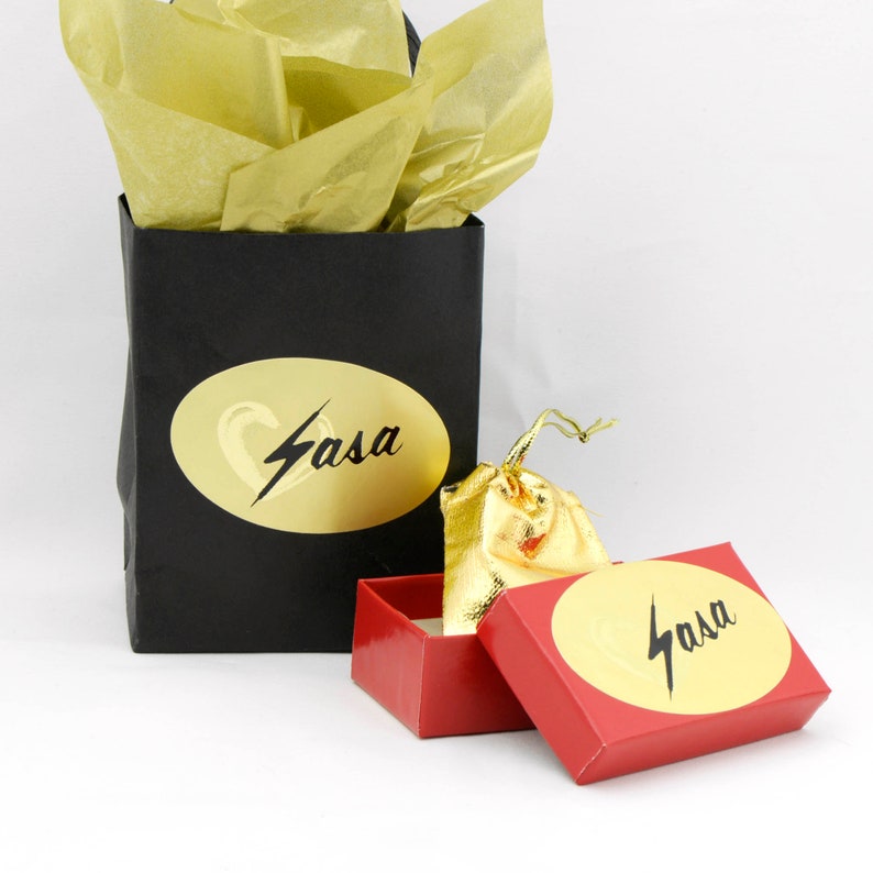 May include: A black gift bag with gold tissue paper, a red box, and a gold drawstring bag. The gift bag and box have a gold oval sticker with the word "Sasa" in black script. The drawstring bag is gold.