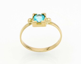 May include: A gold ring with a light blue gemstone set in a square frame. The ring has small gold beads on either side of the gemstone.