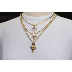 K&ouml;nnte beinhalten: Eine mehrreihige goldene Kettenkette mit drei Kristallanh&auml;ngern. Der untere Anh&auml;nger ist ein runder Kristall mit einem gr&uuml;nen Edelstein-Akzent. Der mittlere Anh&auml;nger ist ein sichelf&ouml;rmiger Kristall. Der obere Anh&auml;nger ist ein dreieckiger Kristall.