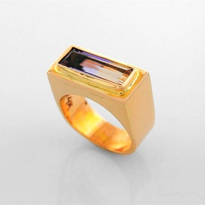 Puede incluir: Un anillo de oro con una piedra preciosa rectangular de amatista engastada en el centro. El anillo tiene una banda cuadrada.