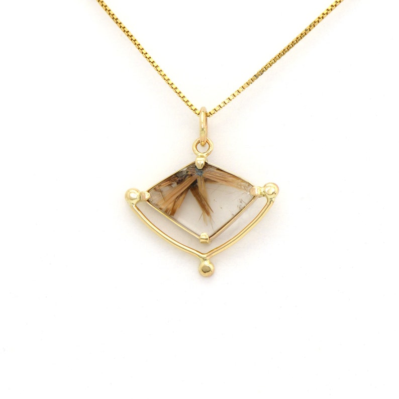 K&ouml;nnte beinhalten: Eine goldene Halskette mit einem Anh&auml;nger aus klarem Quarzkristall mit braunen Einschl&uuml;ssen, gefasst in einem geometrischen Rahmen.