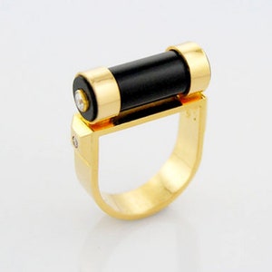 May include: A gold ring with a black onyx stone set in a gold band. The ring has a unique design with the stone set in a cylinder shape.