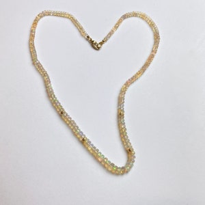 May include: A delicate necklace made of small, white opal beads with gold accents. The necklace is arranged in a heart shape.
