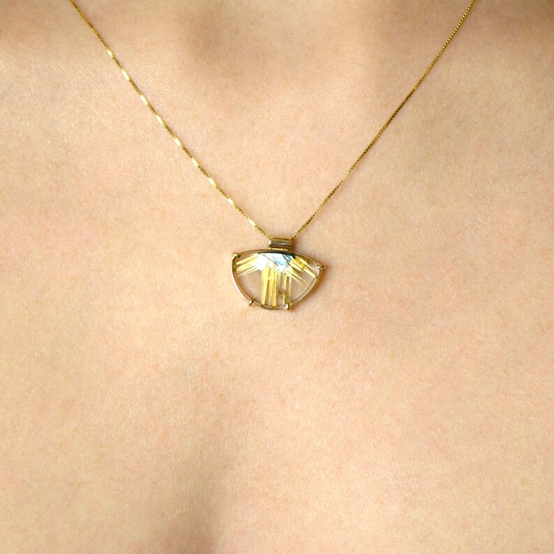 May include: A gold necklace with a pendant featuring a clear, semi-circular stone with gold-colored inclusions. The pendant is suspended from a delicate gold chain, creating a simple yet elegant design. The necklace is shown against a skin-toned background.