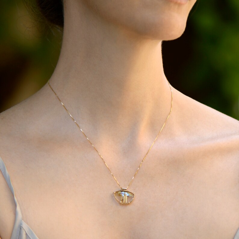 May include: A delicate gold necklace with a pendant featuring a clear, faceted gemstone. The pendant has a unique shape and is suspended from a thin gold chain. The necklace is worn around the neck.