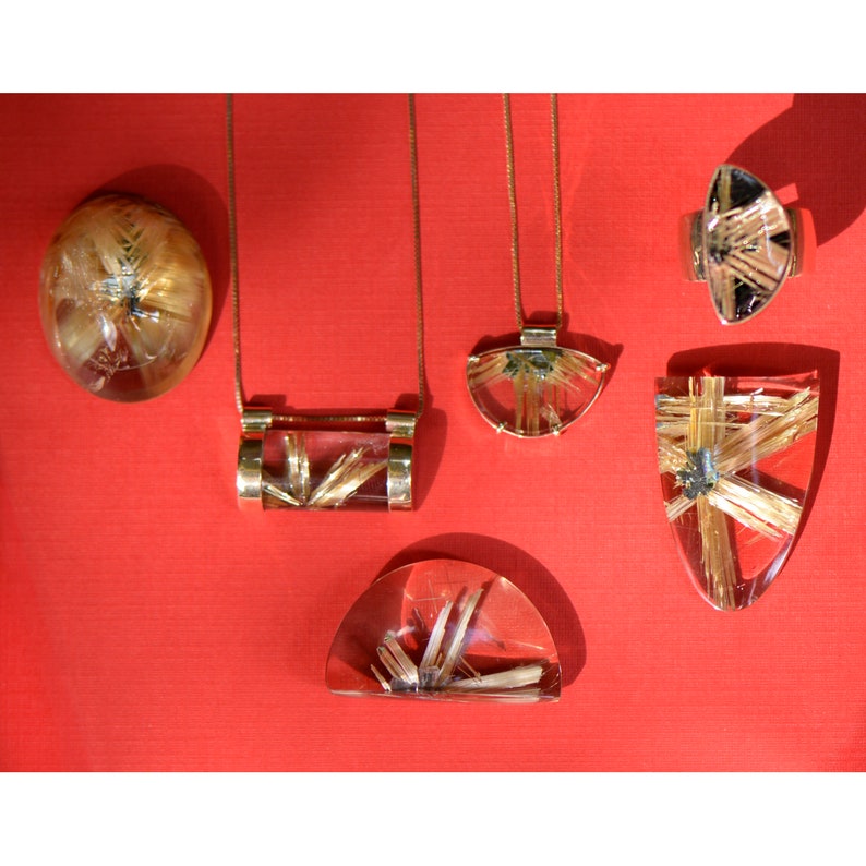 May include: A collection of jewelry pieces featuring clear resin with gold-colored inclusions. The set includes a pendant necklace, a ring, and a pendant. The jewelry is displayed on a red surface.