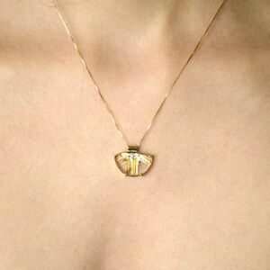 May include: A gold-toned pendant necklace featuring a translucent, fan-shaped stone with gold-colored inclusions. The pendant is suspended from a delicate gold chain. The necklace is displayed against a skin-toned background.