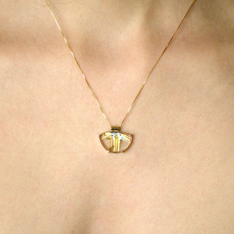 May include: A gold-toned pendant necklace featuring a translucent, fan-shaped stone with gold-colored inclusions. The pendant is suspended from a delicate gold chain. The necklace is displayed against a skin-toned background.