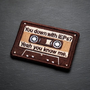 May include: Embroidered patch in the shape of a cassette tape. The patch is brown and tan with the text "You down with IEPs? Yeah you know me." The design includes details like the tape reels and the cassette tape's label.
